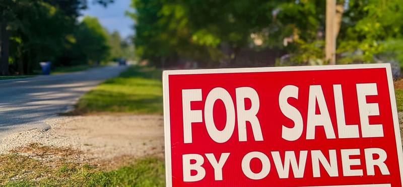 A red FOR SALE BY OWNER sign at the edge of a quiet road