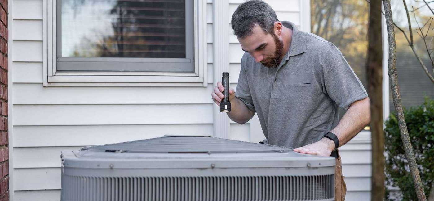 An HVAC technician checking a condenser unit beside a house