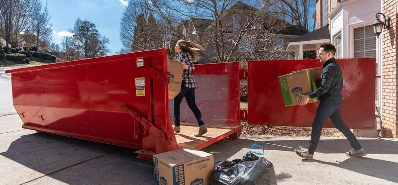 A red roll-off container in a driveway, two people carrying boxes and bags out to it