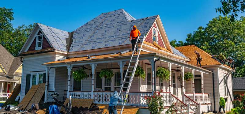 A crew re-roofing a large house with a wraparound porch, ladder against the eaves